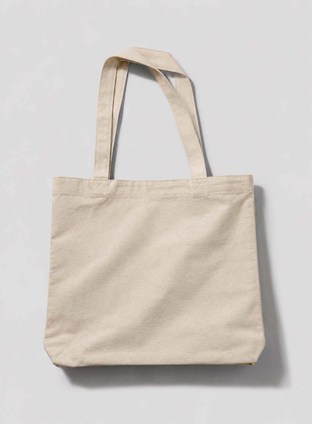 A plain canvas tote bag in a light beige or cream color, shown from a front-facing perspective against a neutral light gray background.