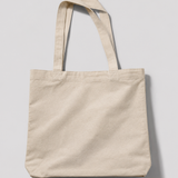 A plain canvas tote bag in a light beige or cream color, shown from a front-facing perspective against a neutral light gray background.